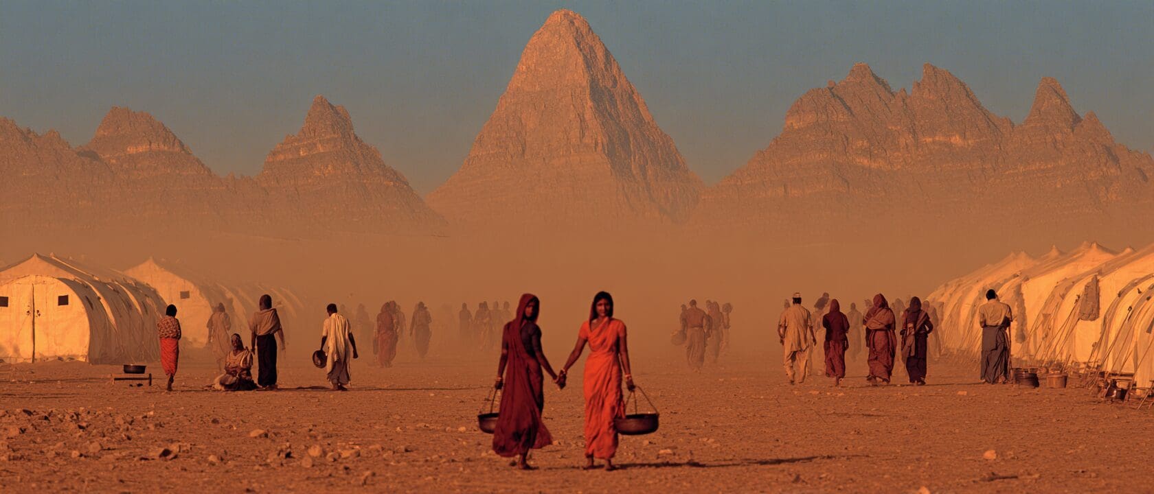 Two women in red and orange saris walk hand in hand across a dusty plain carrying water vessels, with the mountains of Kurukshetra behind them — evoking the ethic of care on the Bhagavad Gita's battlefield